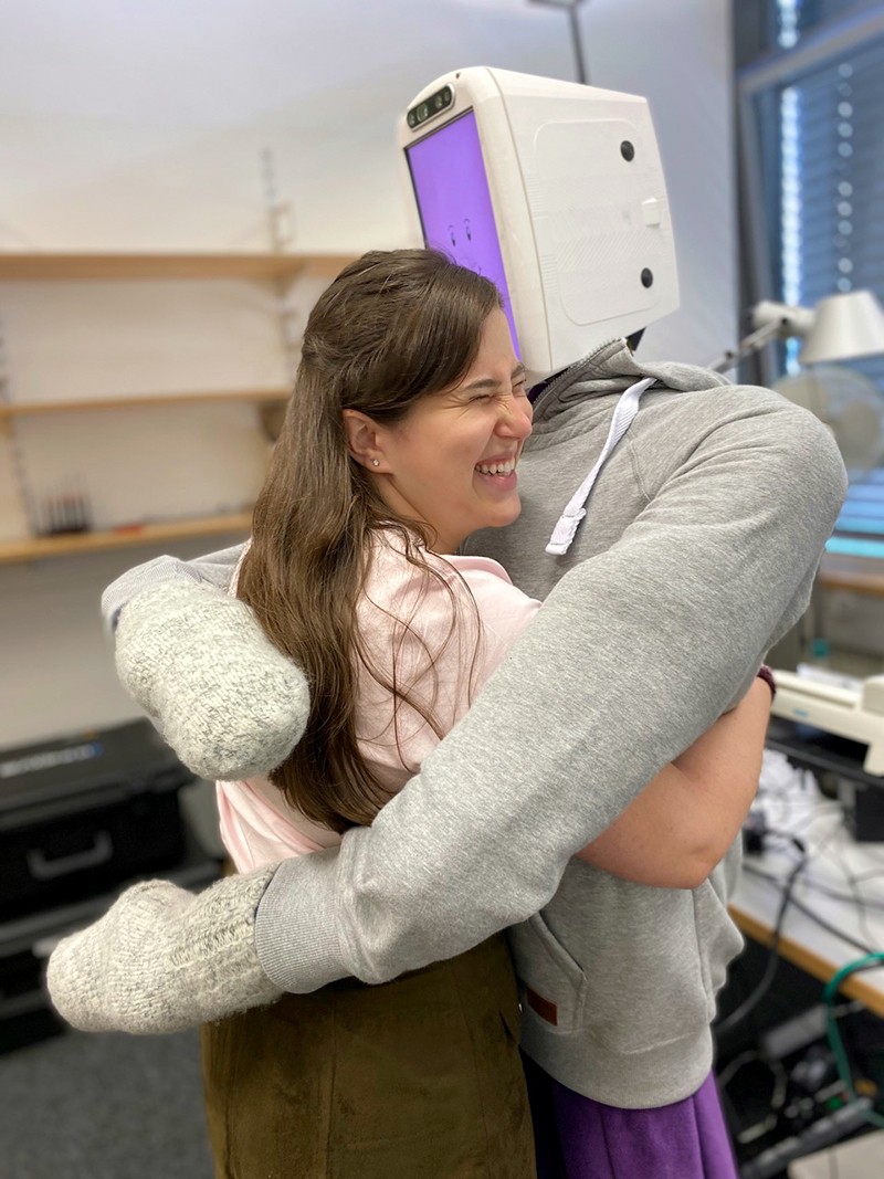 A robot with a computer head and wearing a hoodie hugs a woman who is laughing.