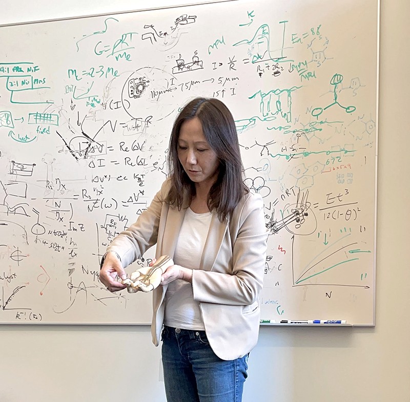 Zhenan Bao in front of white board, pressing on the tip of a finger of an artificial hand.