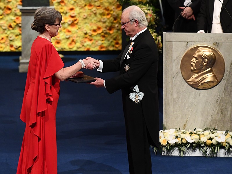 A woman in a red dress shakes hands with and receives a prize from a man in a dark suit.