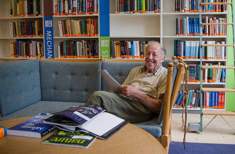 Portrait of Ben Mottelson sitting on a sofa with a bookshelf behind him
