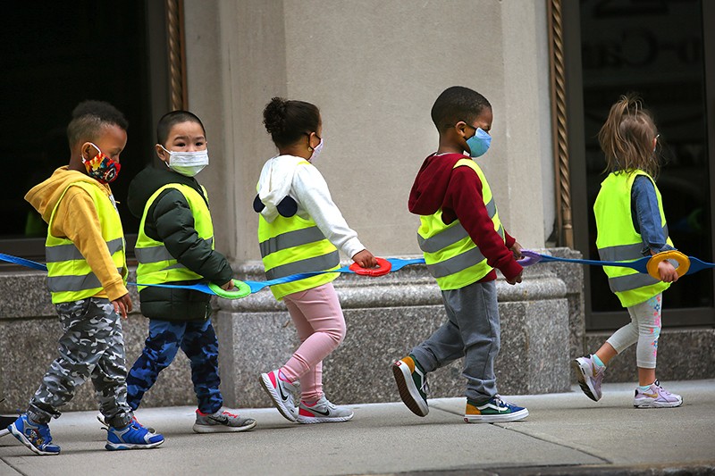 Los niños de una clase de preescolar usan chalecos y máscaras y se sujetan de una correa mientras caminan por una calle de Boston.