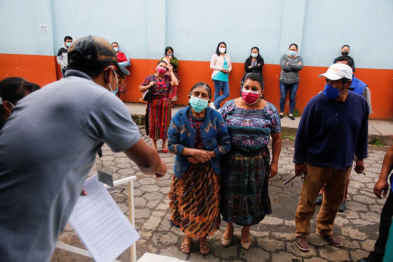 People in masks and colorful clothes listen to a man holding a piece of paper on the street.