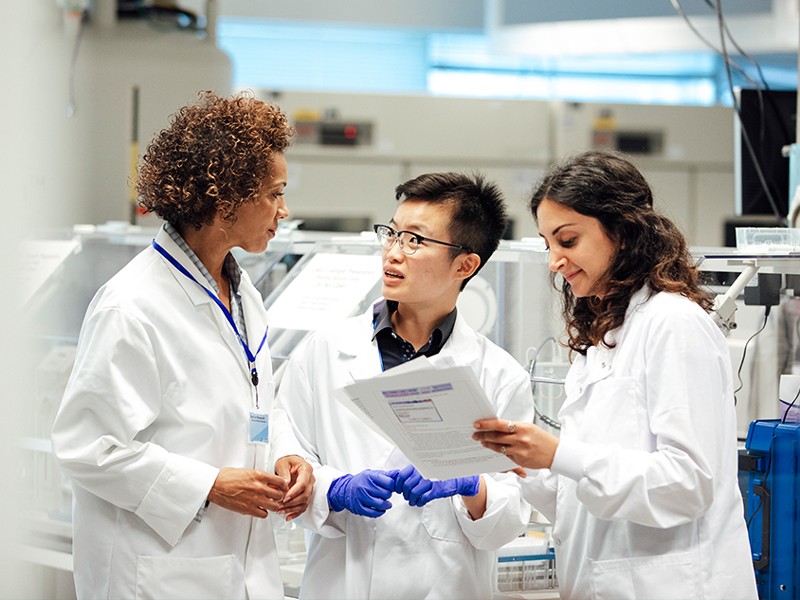 Diverse mix of technicians, dressed in lab coats, discuss work procedure in a laboratory.