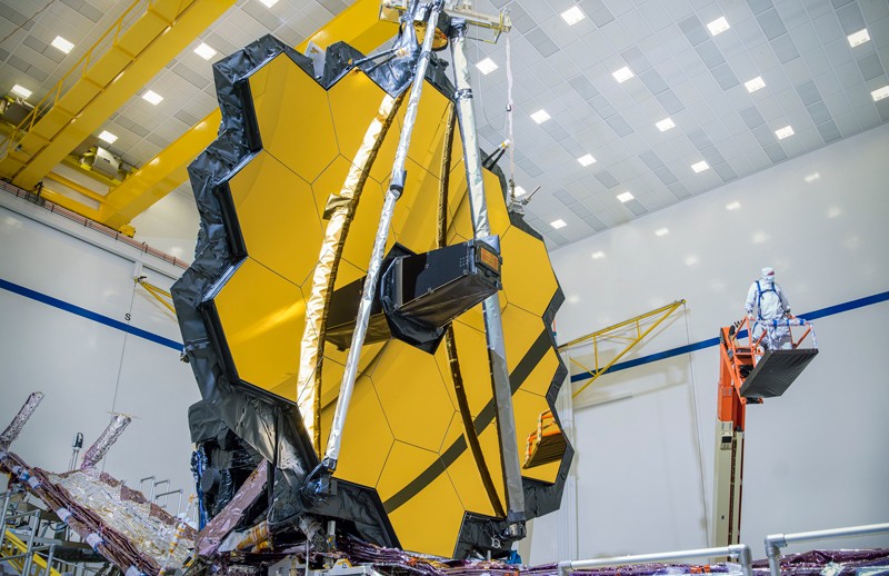 An engineer in a clean room suit stands on a pod next to the primary mirror of the James Webb Telescope