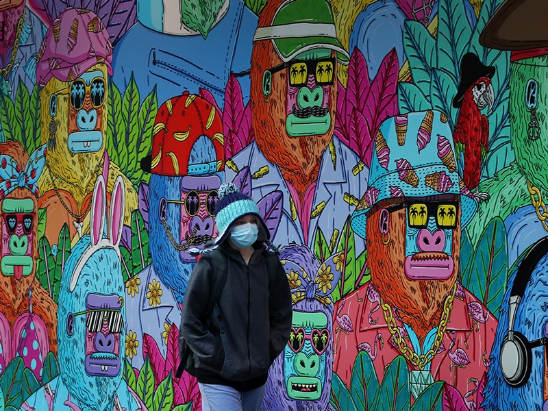 A person in a mask and hat, walking in front of a wall covered in colourful art.