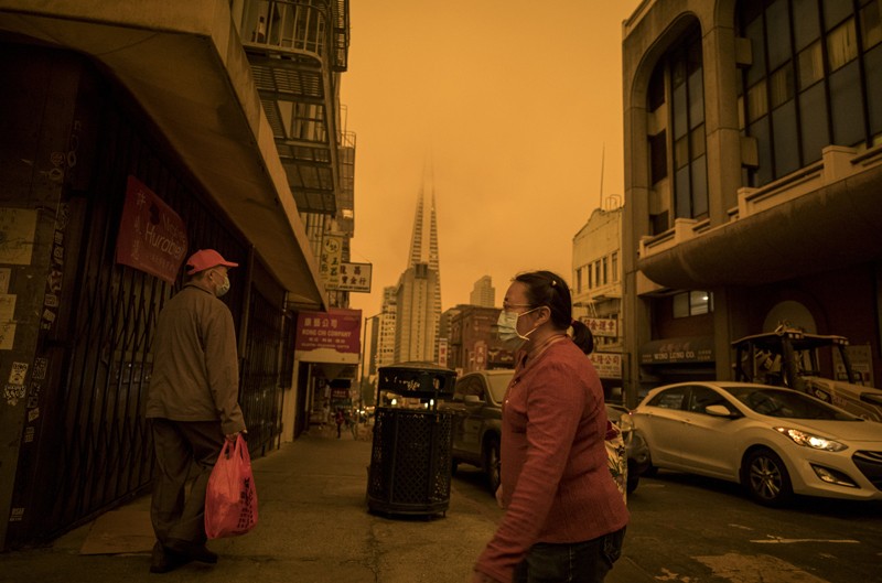 People wearing protective face masks walk along a street while the sky glows orange