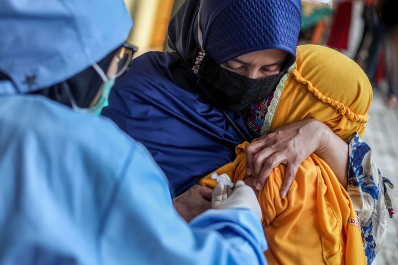A women in a face covering comforts a child as a health worker injects them with a tetanus diphtheria vaccine