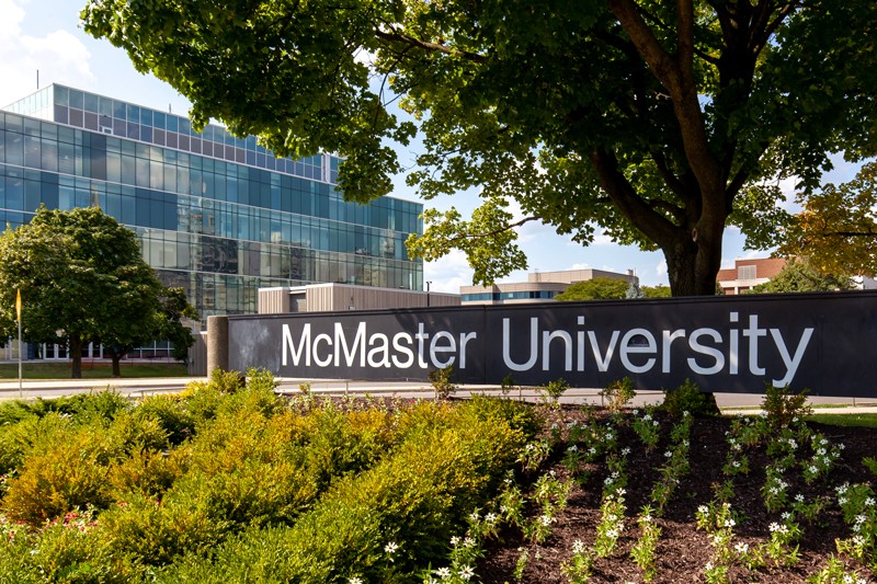 En el campus se ve un letrero de la Universidad McMaster con un edificio de vidrio al fondo