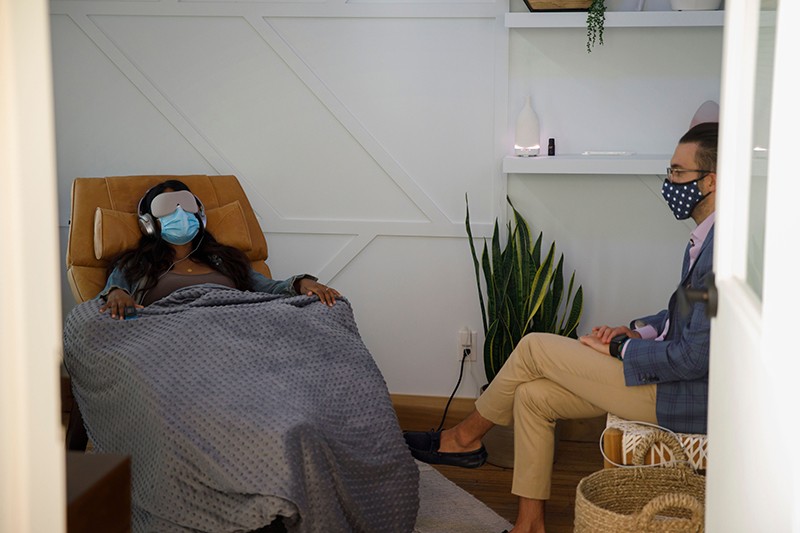 A woman wearing headphones, eyemask and a facemask lies in a recliner under a blanket. A doctor sits to her right.