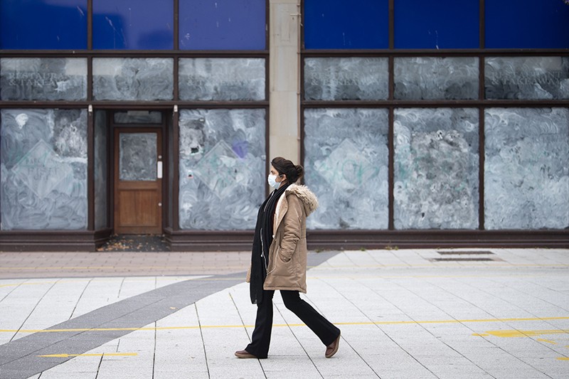 A woman in a coat and facemask walks past a row of closed shops with their windows painted white