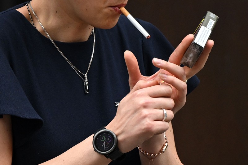 A young woman holds a cigarette packet while lighting a cigarette