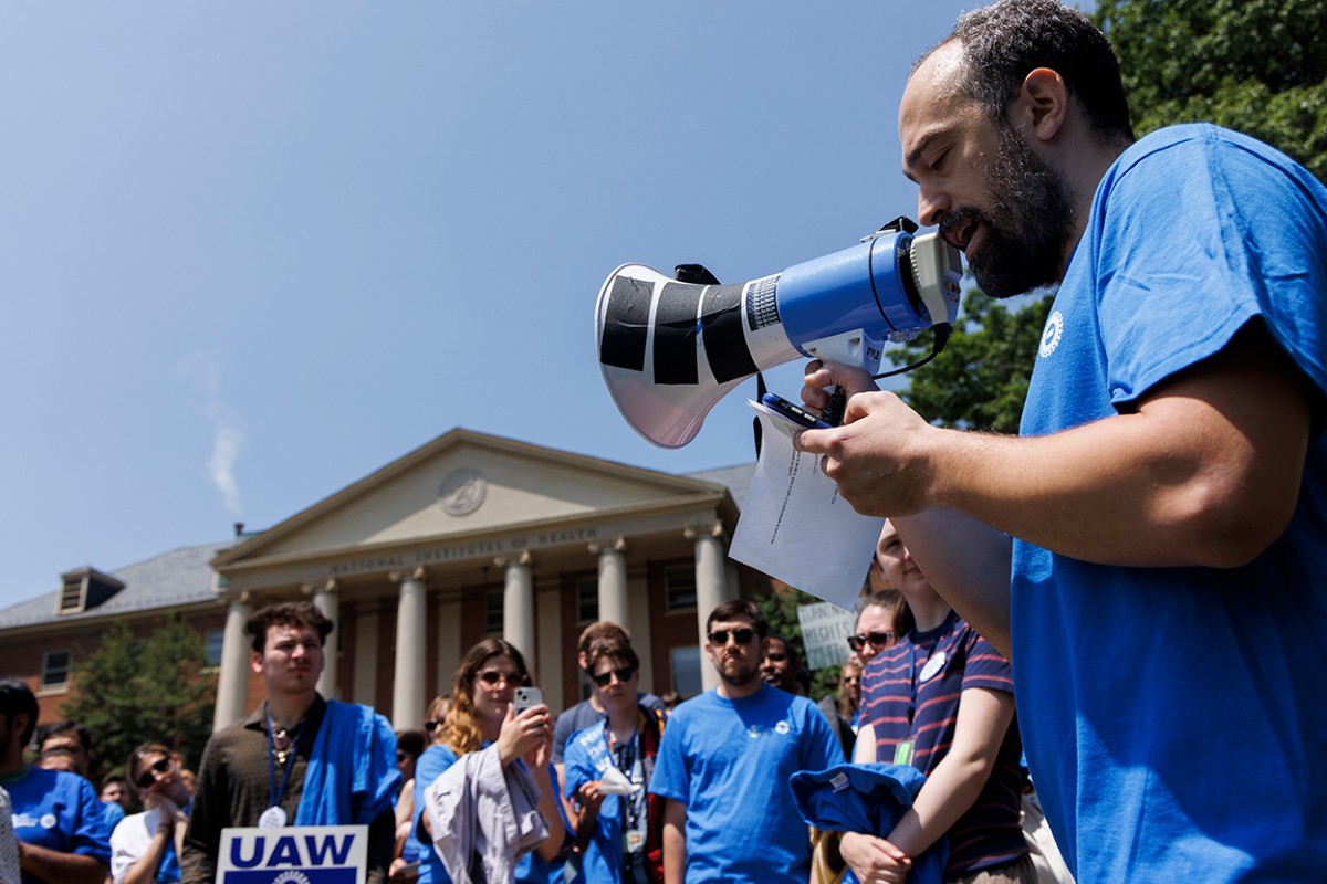 NIH research fellow speaking through a megaphone at a Fellows organizing rally on the NIH Bethesda campus, with Building 1 behind him.