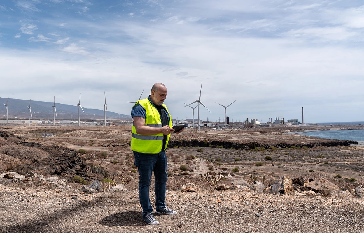 Guillermo Galván García in high-vis jacket holding a tablet standing among wind turbines near the coast with a bright sky above him.