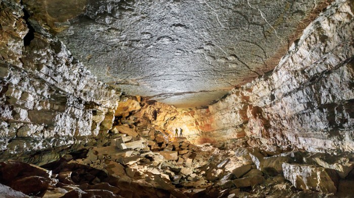 Long, wide cave with raised patches across ceiling and two people in the distance in silhouette against a light.