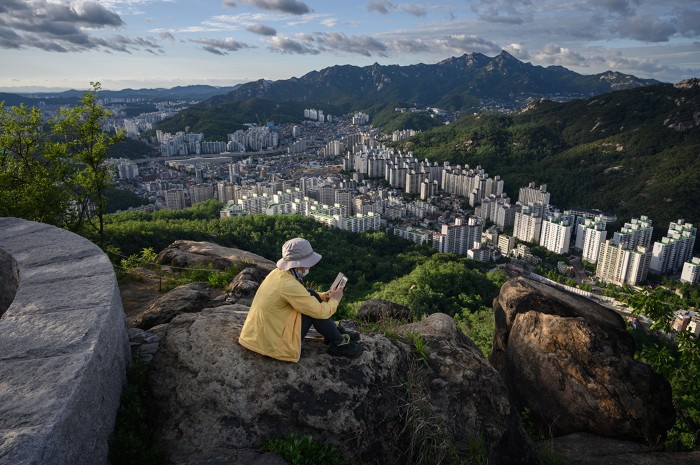 A woman sits looking at her phone on a hillside above the Seoul city skyline