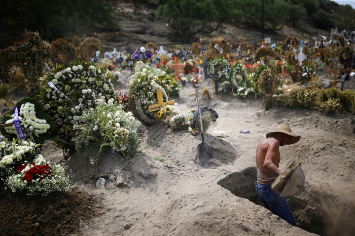 A cemetery worker digging a grave in a site surrounded by newly dug graves covered in memorials and flowers