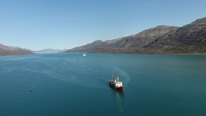 Aerial photograph of a ship in a fjord, with an iceberg in the distance.