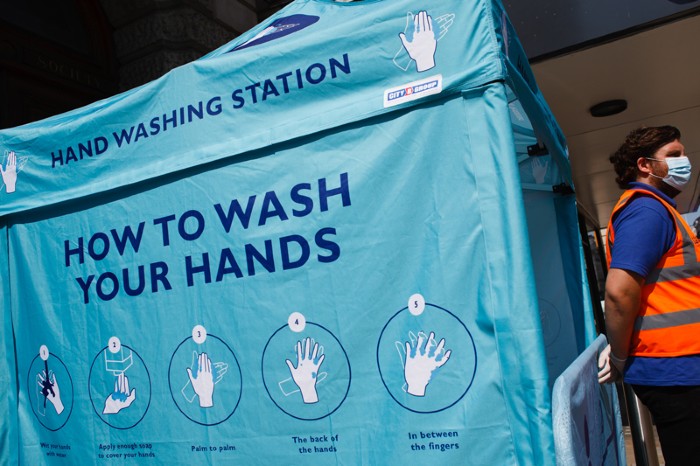 A social-distancing ambassador wearing a face mask mans a hand-washing station.