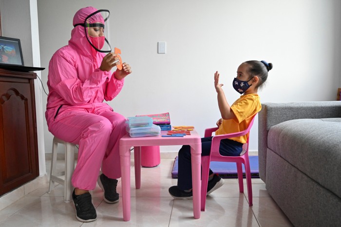 A teacher wearing a bright pink biosafety suit and face shield teaches numbers to a young girl at home