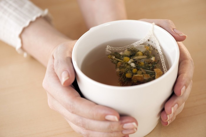 A woman's hands holding a white cup with chamomile tea in a teabag steeping in water.