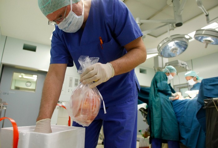 A surgeon puts a plastic bag containing a kidney into a box while a patient is being operated on in the background