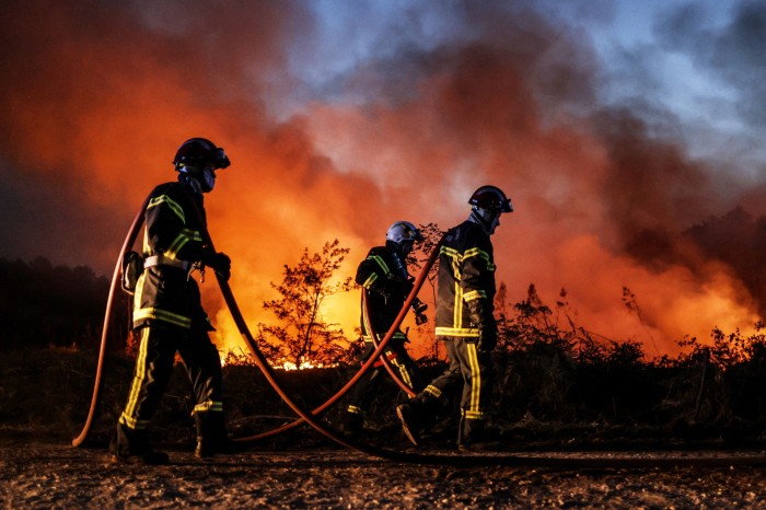 Three firefighters carry a hose pipe past a wildfire burning in France during a heatwave