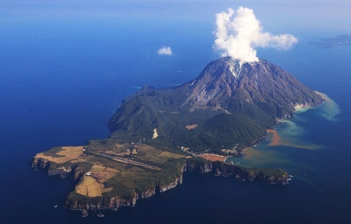 An aerial photo of a small island. At the far end is a mountain emitting gas and smoke.