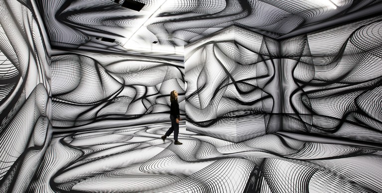 A white woman dressed in black stands in a room with the walls, ceiling and floor covered in swirling black and white patterns.