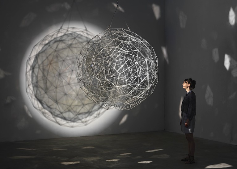A woman in a darkened room looks at a ball of steel wire casting polyhedral shadows.