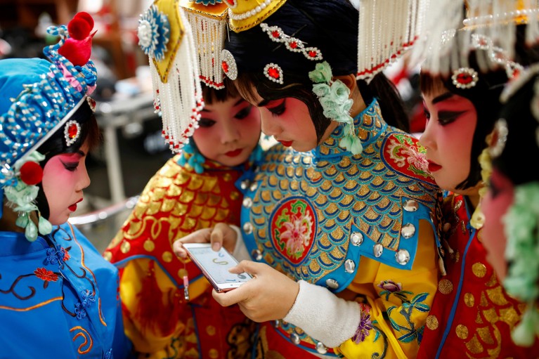 A teenage participant plays a game on her phone as others watch during a break in a traditional Chinese opera competition