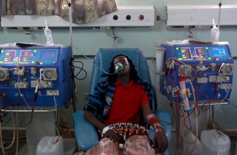 A patient wearing a ventilator mask sits in a chair between two large blue dialysis machines covered in tubes and wires