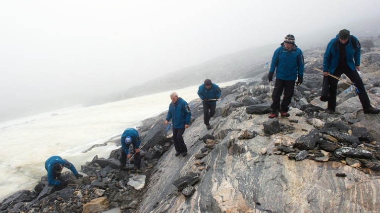 Six people in blue coats use sticks to search for objects among rocks next to an expanse of ice.