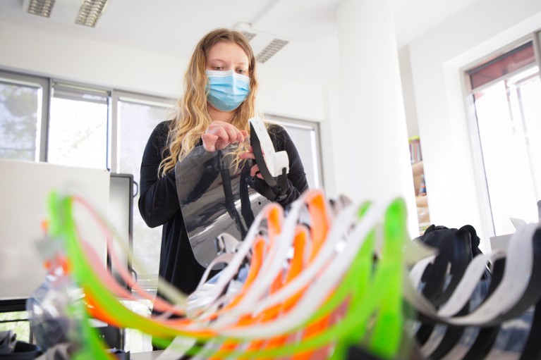 A young woman wearing a surgical mask assembles a 3D printed protective mask with finished masks piled on a table in front