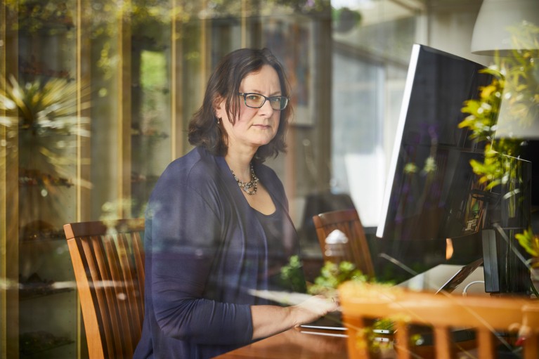 Elisabeth Bik poses for a portrait taken through glass reflecting a garden showing her sitting at a table with a large monitor