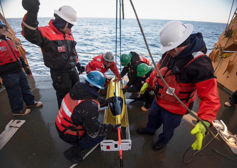A ships crew members in hard hats prepare to hoist a yellow, rocket-shaped sea glider