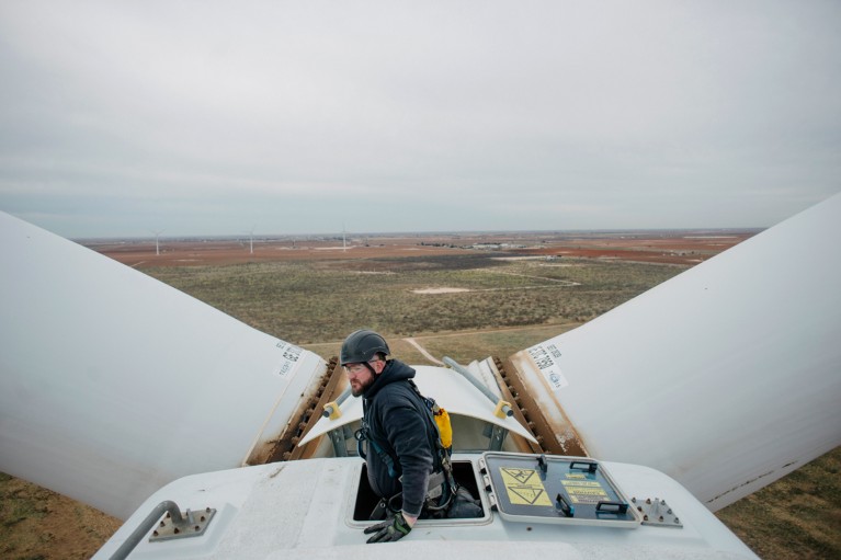 A man in hard hat emerging from the top of a wind turbine surrounded by fields and turbines in the distance