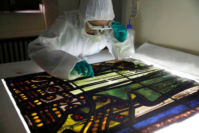 Claudine Loisel, wearing full body covering and gloves, peers closely at a back-lit stained-glass window on a table in a lab