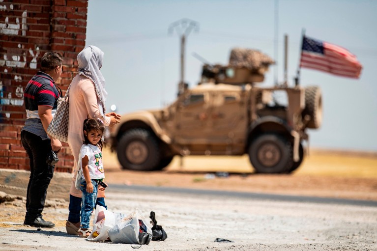 A family with a young girl and bags stand in a road in Hasakeh province, Syria watching a military car with an American flag