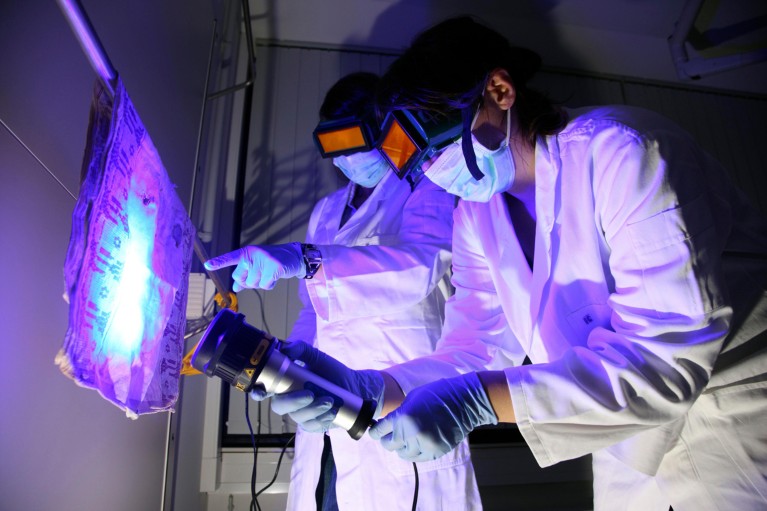 Two people wearing orange tinted goggles, lab coats and gloves use a UV torch to find traces of DNA on a piece of fabric