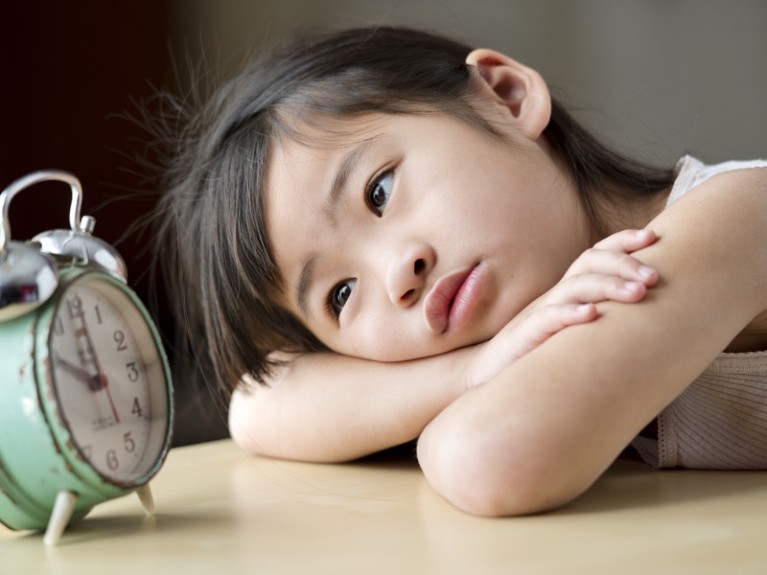 Portrait of a little girl sitting with her head on her arms, staring at an old clock.