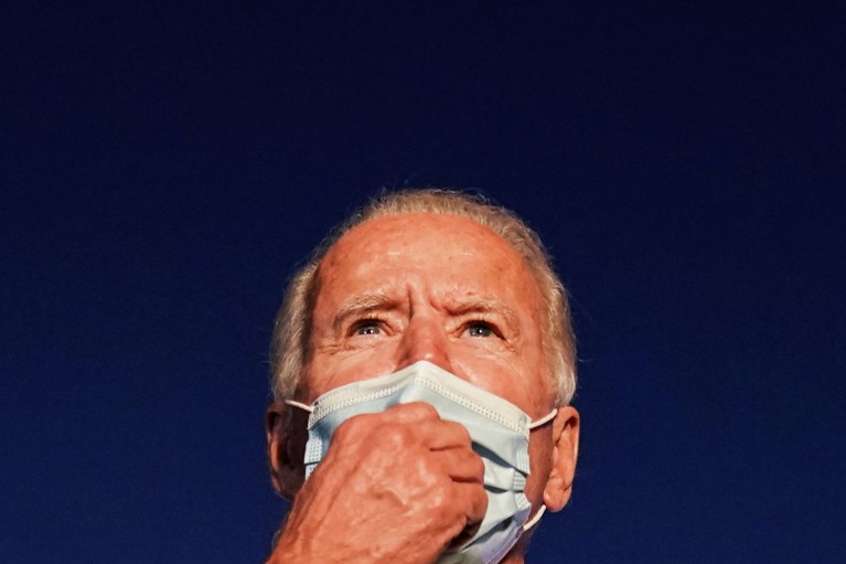 Close up of Joe Biden wearing a face mask looking into the distance on a deep blue background