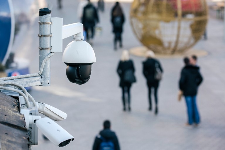 A spherical security camera overlooks a pedestrianised space with people walking past a large globe-shaped sculpture