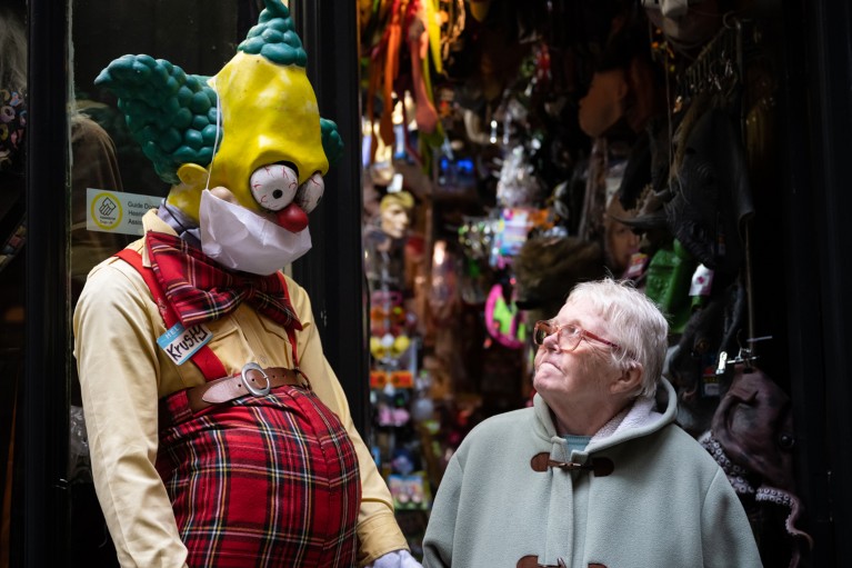 An elderly woman looks up at a large model of a clown character in a surgical face mask outside a fancy dress shop in Cardiff