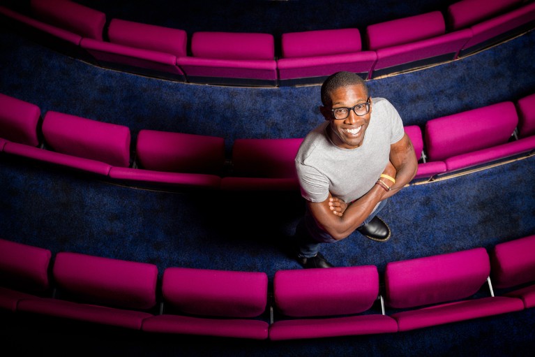 A portrait of Chris Jackson looking. up to camera whilst standing between curved rows of pink folded seats in a lecture theatre