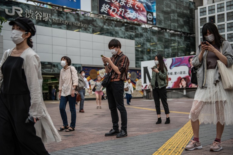 People standing apart and wearing face masks look at their phones while waiting to cross the road in Tokyo
