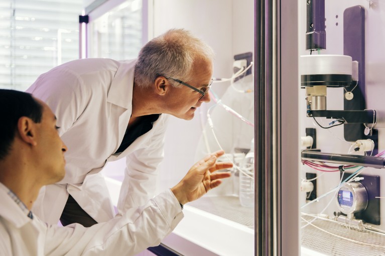 Two workers in white lab coats inspect a large electronic system behind a glass screen