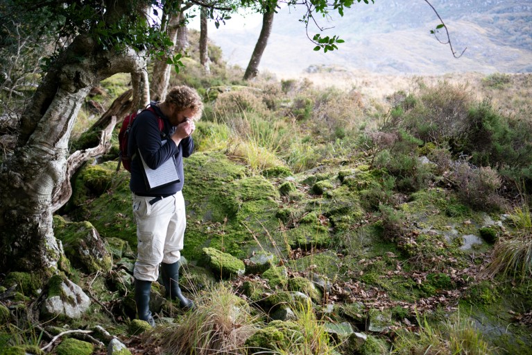 Rory Hodd stands amongst trees, ferns and rocks looking through a small magnifying glass at a sample of moss