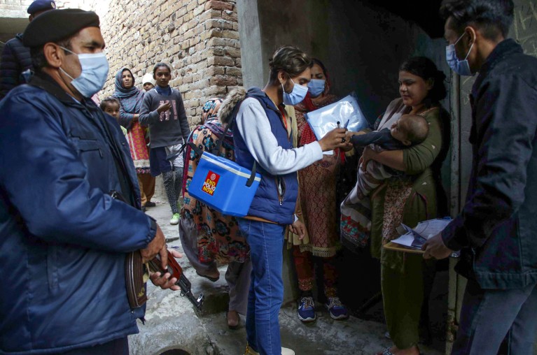 在巴基斯坦，一名男性卫生工作者拿着一个凉爽的盒子给婴儿注射小儿麻痹症疫苗，武装警察在一旁守卫