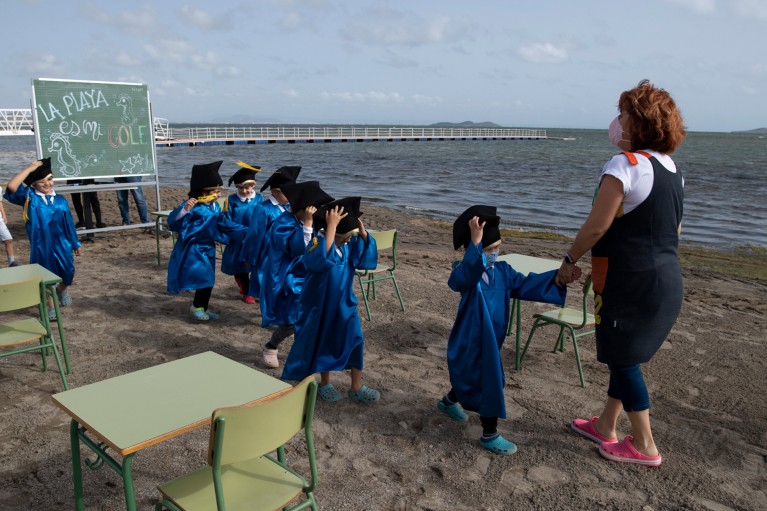 Young school pupils wearing gowns and mortarboards are led by a teacher during a graduation ceremony on a beach in Spain