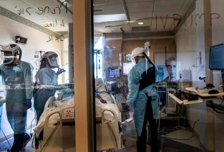 A doctor's face on a screen is reflected in a window in an intensive care unit with medical workers treating a COVID-19 patient